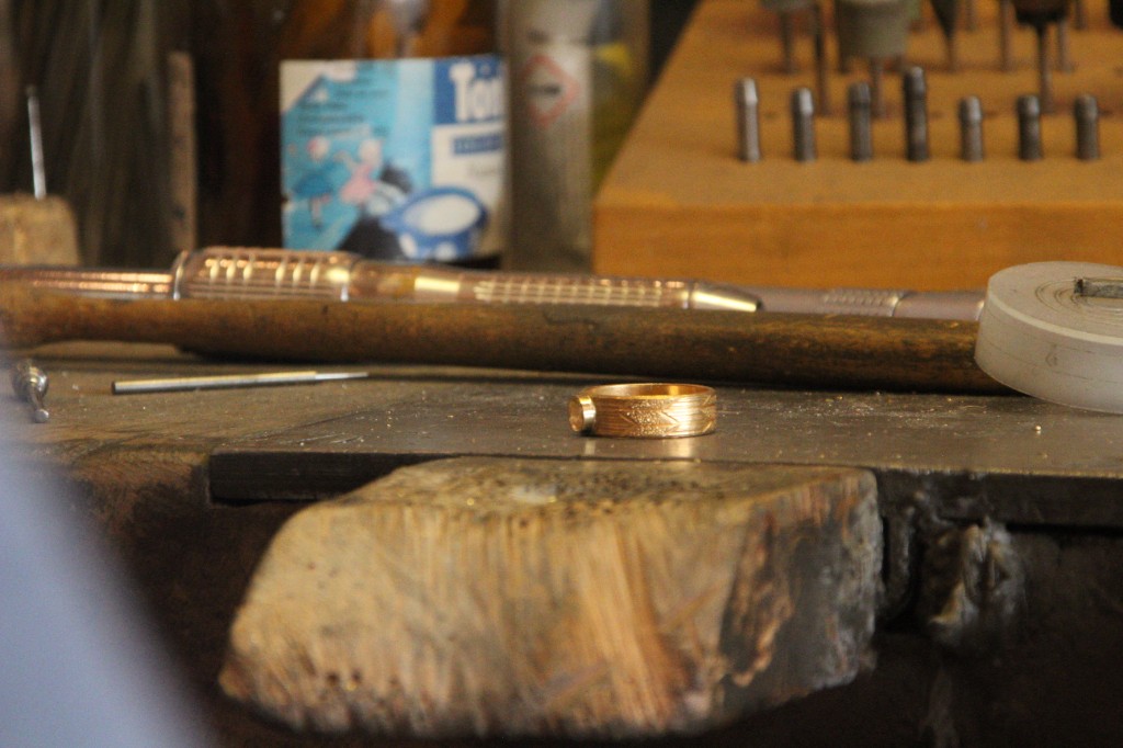 Ein Goldring mit feiner Gravur steht auf der Werkbank, dahinter Werkzeug und Bohrer
